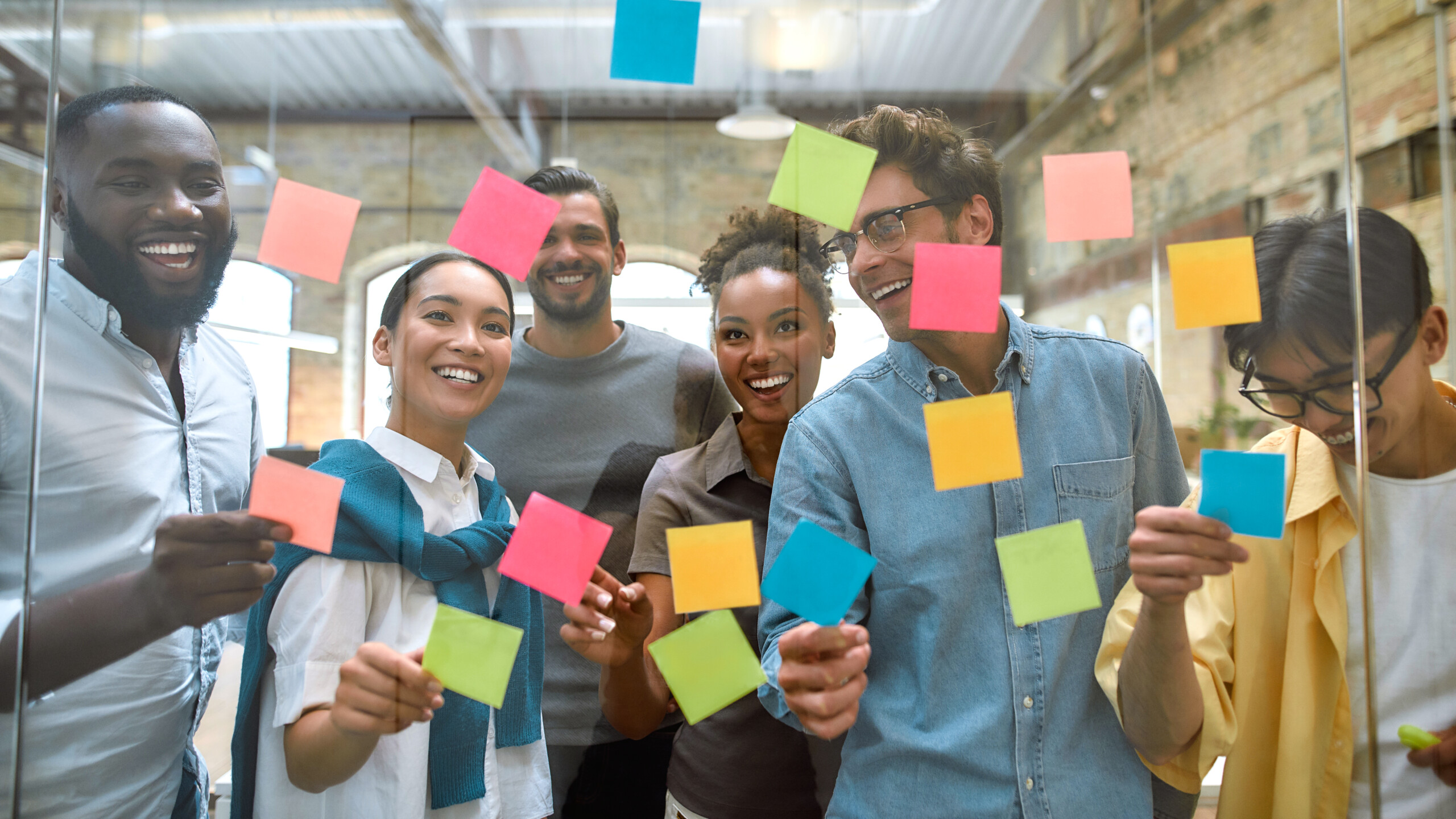 Working on business project together. Group of young and positive coworkers putting colorful sticky notes on a glass window in the creative office